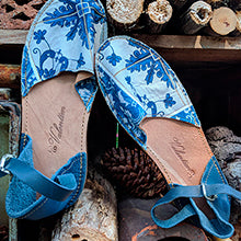 The Valentims Ladies Espadrille Sandals by Iberica feature a blue and white pattern and ankle straps, displayed on stacked firewood with pinecones in a rustic setting.