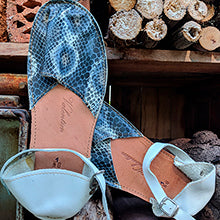 The Valentims Ladies Espadrille Sandals by Iberica feature blue and white snakeskin-patterned uppers and white ankle straps. Handmade in Portugal, these leather sandals are displayed against stacked firewood.
