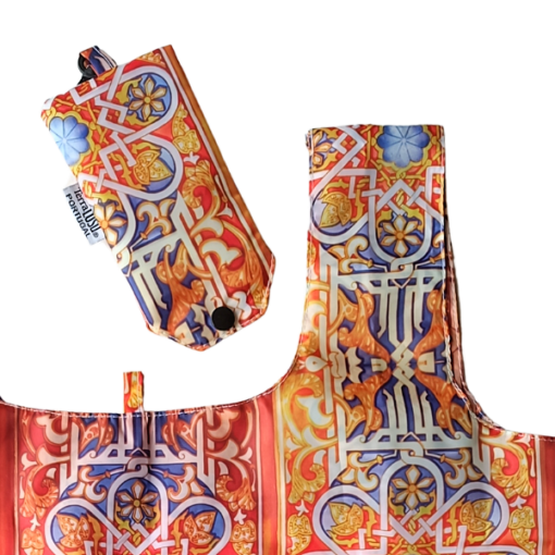 The Terra Lusa Stained Glass Eco Shopper, featuring a vibrant red, blue, yellow, and orange pattern, is shown partially unfolded beside its matching compact pouch.