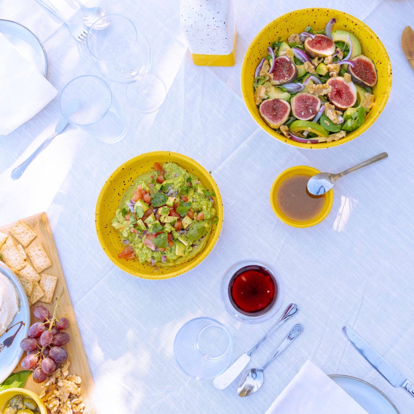 A vibrant outdoor table displays yellow salad bowls with figs and guacamole, a glass of red drink, and the Alfar Tierra Cocida Ceramic Cheese and Vegetable Grater Plate/Nuevo Canela beside crackers, walnuts, and grapes on a white tablecloth.