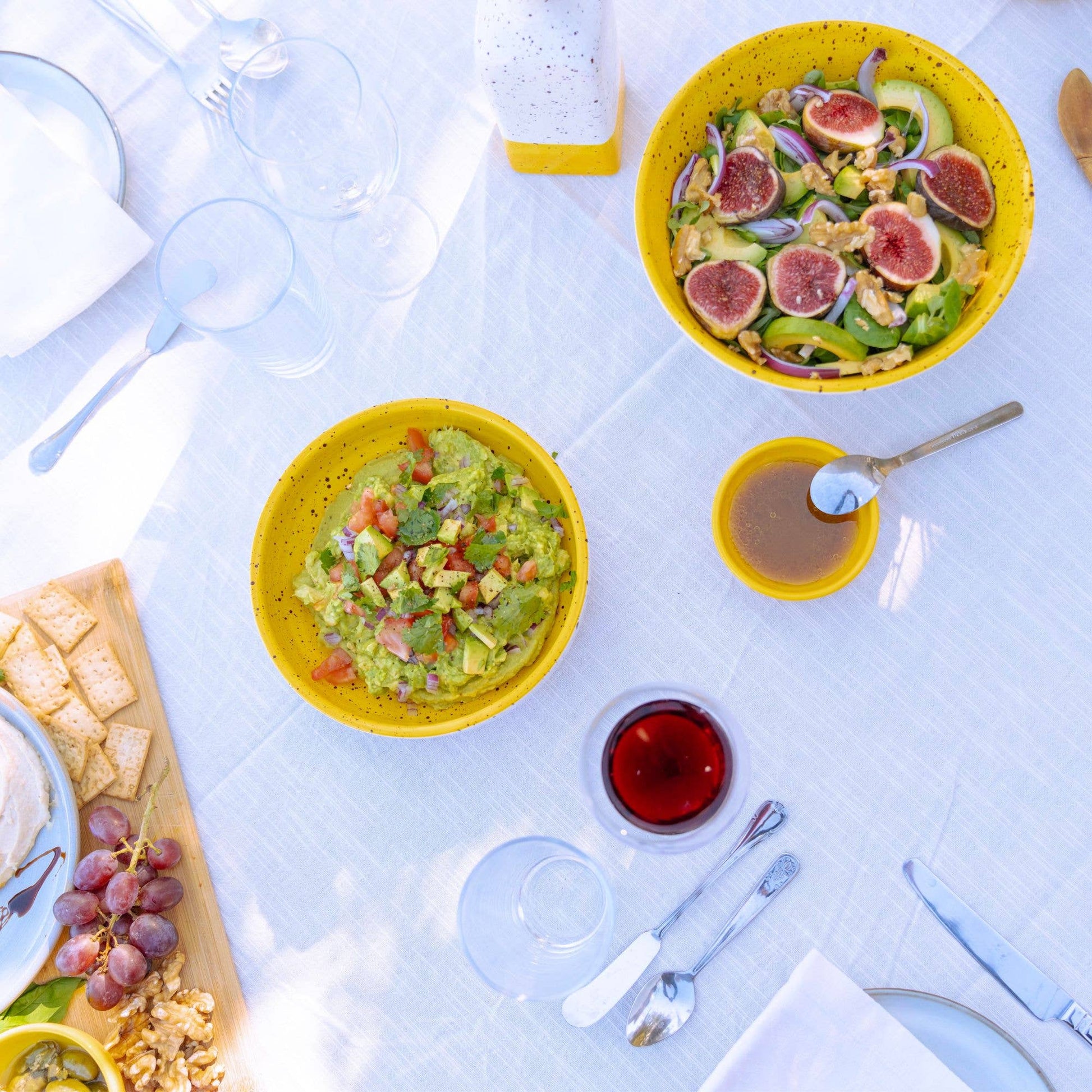 A vibrant outdoor table displays yellow salad bowls with figs and guacamole, a glass of red drink, and the Alfar Tierra Cocida Ceramic Cheese and Vegetable Grater Plate/Nuevo Canela beside crackers, walnuts, and grapes on a white tablecloth.