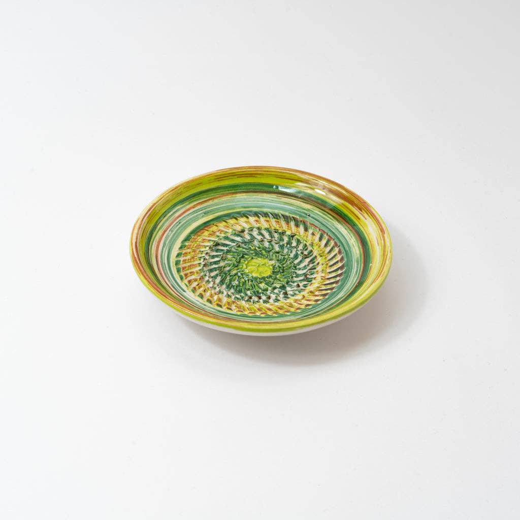 The Alfar Tierra Cocida Ceramic Cheese and Vegetable Grater Plate/Verde Amazo is a handmade, shallow round bowl from Spain with a vibrant green, yellow, and brown spiral and geometric pattern on white.