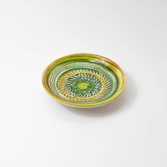 The Alfar Tierra Cocida Ceramic Cheese and Vegetable Grater Plate/Verde Amazo is a handmade, shallow round bowl from Spain with a vibrant green, yellow, and brown spiral and geometric pattern on white.