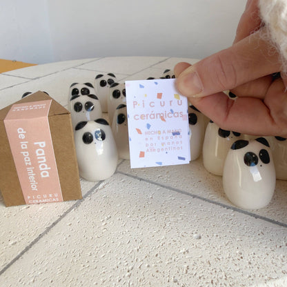 A hand displays a Panda Amulet of Inner Peace – Handmade Lucky Charm Gift by Picuru ceramicas, alongside a branded card and box. These unique items sit on a white textured surface—perfect for gifting tranquility and luck.