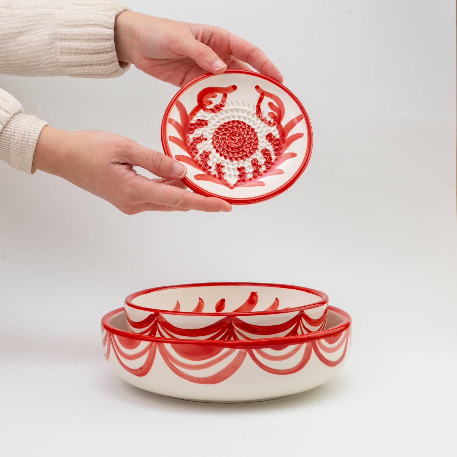 Hands hold the Alfar Tierra Cocida Small Garlic Grater Ceramic Plate – Sevilla above two matching red-patterned nested bowls, all set against a plain white background.