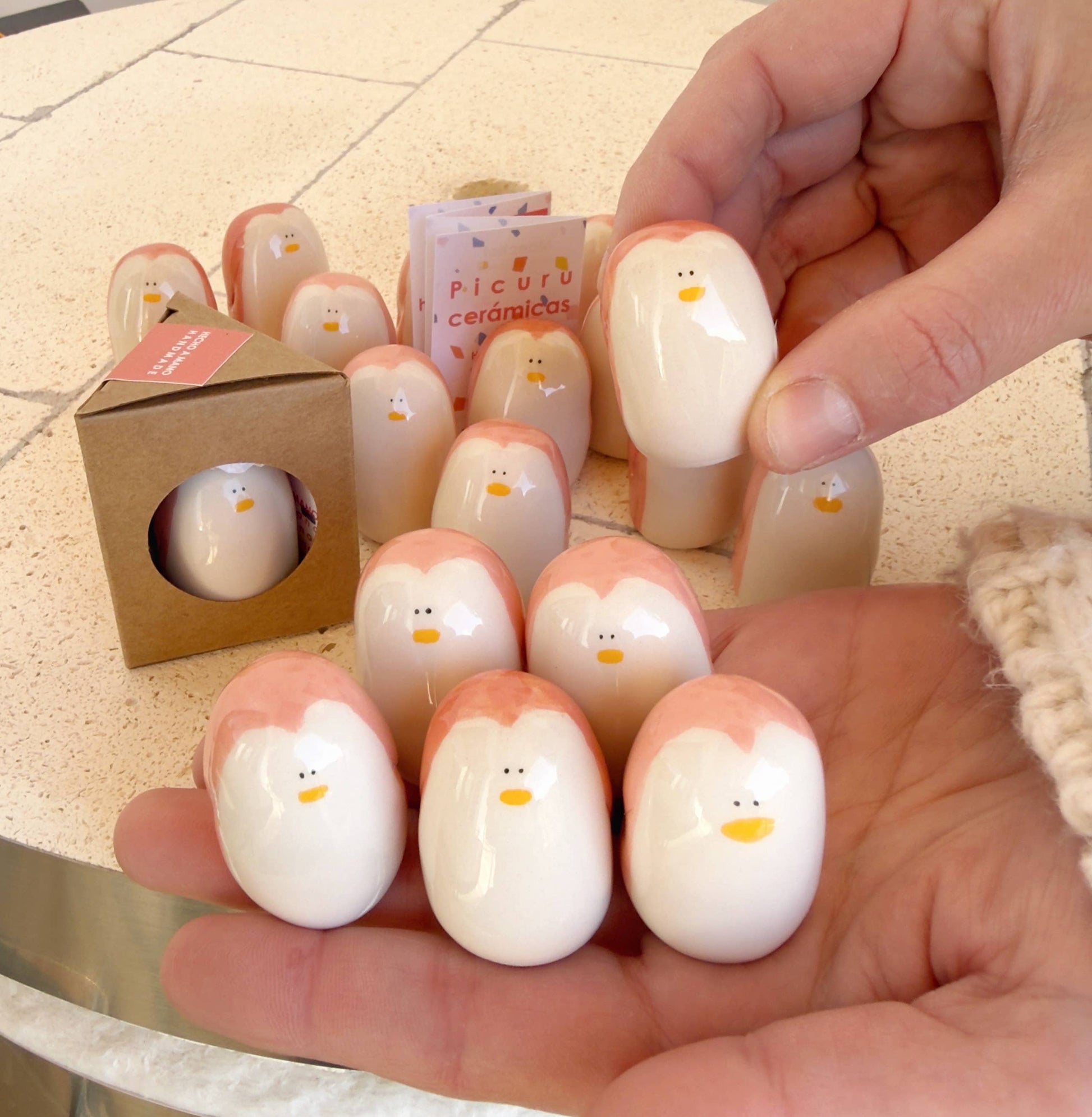 Two hands hold several small, smooth Penguin Amulet of Presence—Handmade Symbols of Balance & Resilience by Picuru ceramicas, featuring light pink tops. More amulets are displayed on a table and in a box labeled Picuru ceramicas.