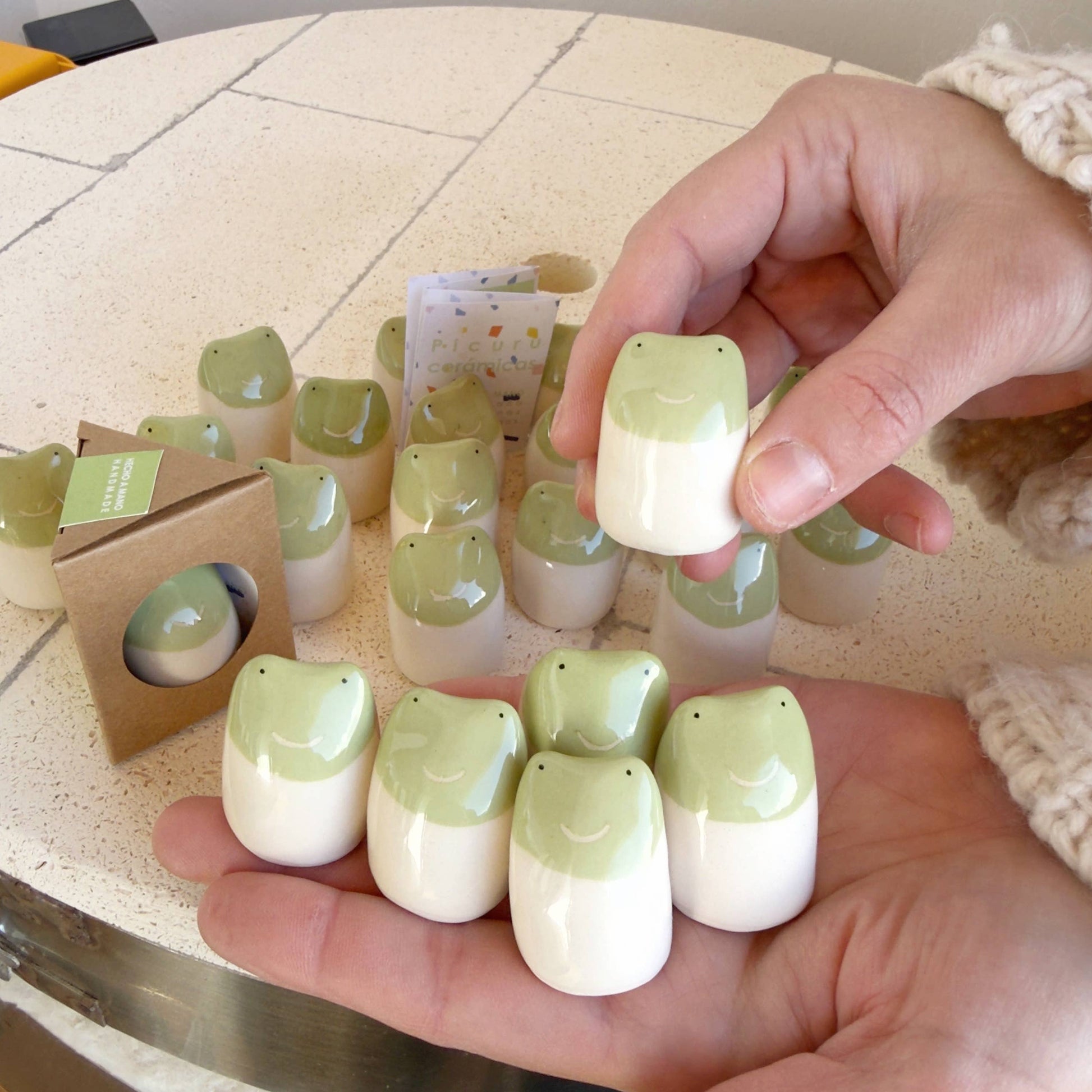 A person holds several Little Frog Amulet of Adventures figurines by Picuru ceramicas—handmade green and white frog charms, symbols of joy and new experiences, with more identical amulets and packaging seen on a table nearby.