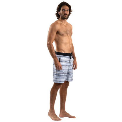 A shirtless man with medium-length brown hair stands barefoot against a plain white background, wearing Futah’s Cacela Blue Boardshorts featuring a drawstring waist and patterned design.