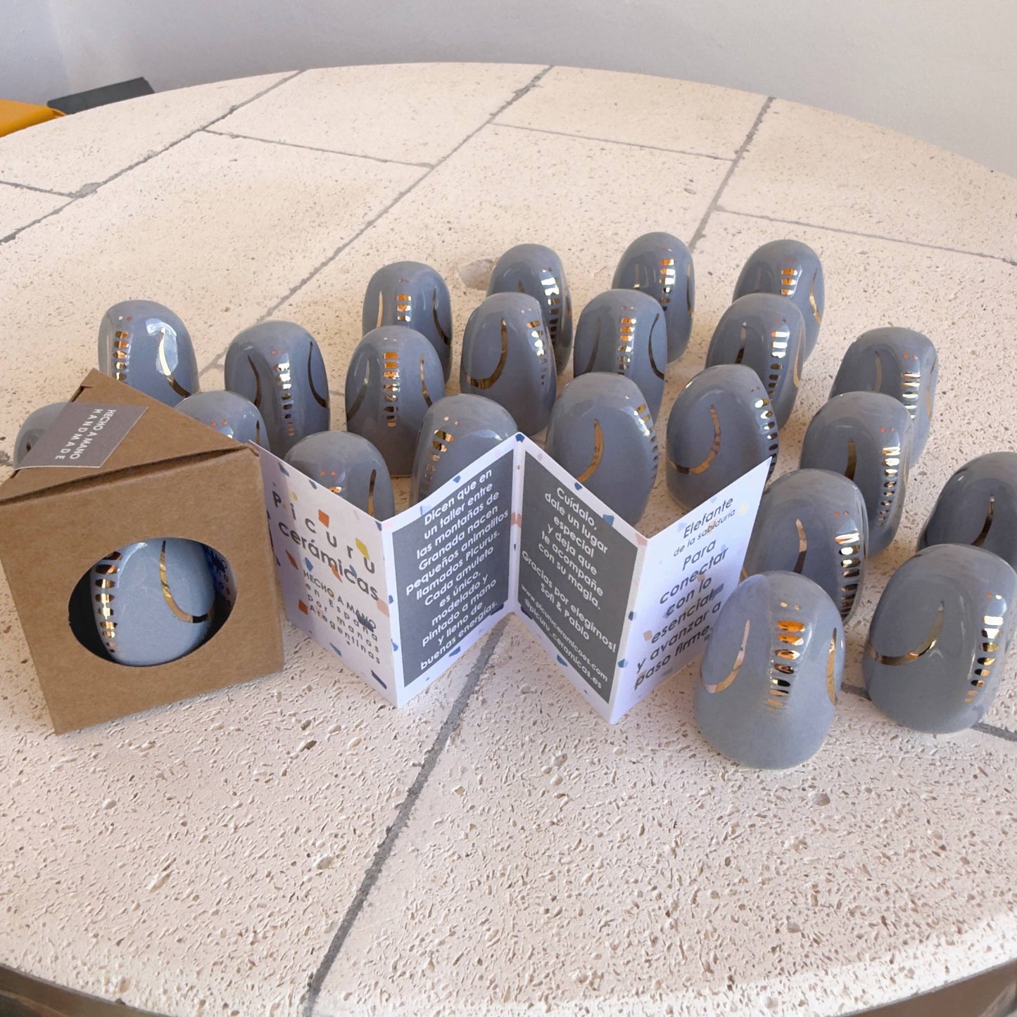 A set of smooth, gray Amule Elephant of Wisdom "Gold" 5 Languages talismans by Picuru ceramicas with gold accents is displayed on a round tiled table, beside an open booklet and a box containing another talisman.