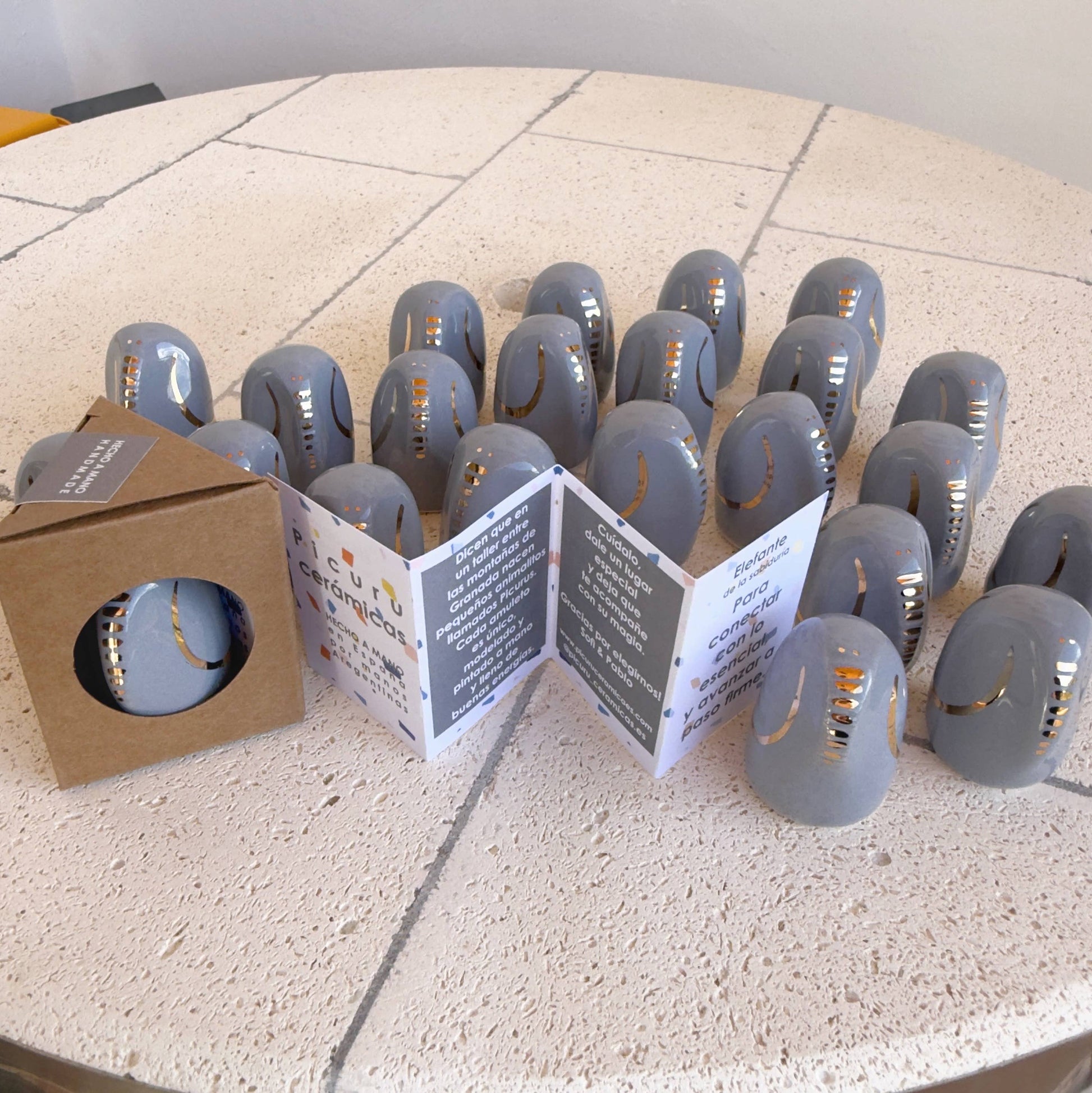 A set of smooth, gray Amule Elephant of Wisdom "Gold" 5 Languages talismans by Picuru ceramicas with gold accents is displayed on a round tiled table, beside an open booklet and a box containing another talisman.