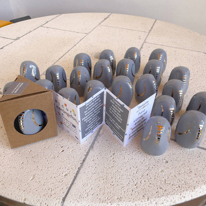 A set of smooth, gray Amule Elephant of Wisdom "Gold" 5 Languages talismans by Picuru ceramicas with gold accents is displayed on a round tiled table, beside an open booklet and a box containing another talisman.