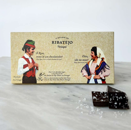 A box of O Melhor do Ribatejo’s Dark Chocolate with Flor de Sal from Portugal rests on marble, adorned with vintage-style art of a traditionally dressed couple and showcasing pieces of dark chocolate and sea salt.