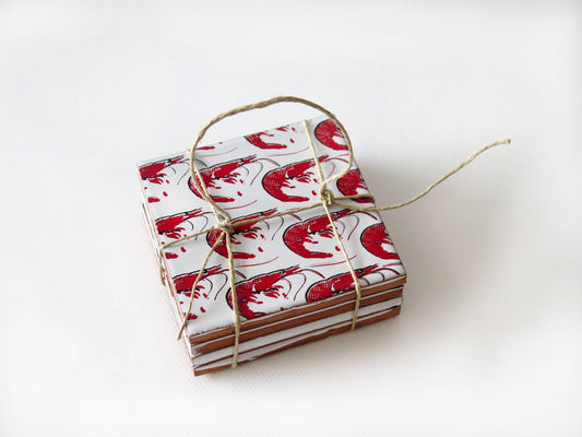 A small stack of Bussoga Gamba Design Ceramic Coasters, featuring red and black shrimp illustrations and tied with brown twine, sits on a white surface.