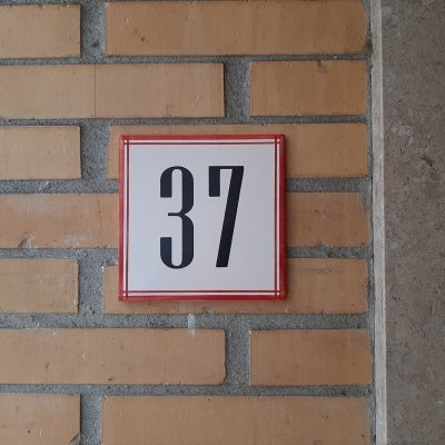A Handpainted Ceramic Door Number by TilePassion, styled like custom Portuguese tiles, displays the number 37 in bold black on a white square with a thin red border, mounted on a yellow brick wall.