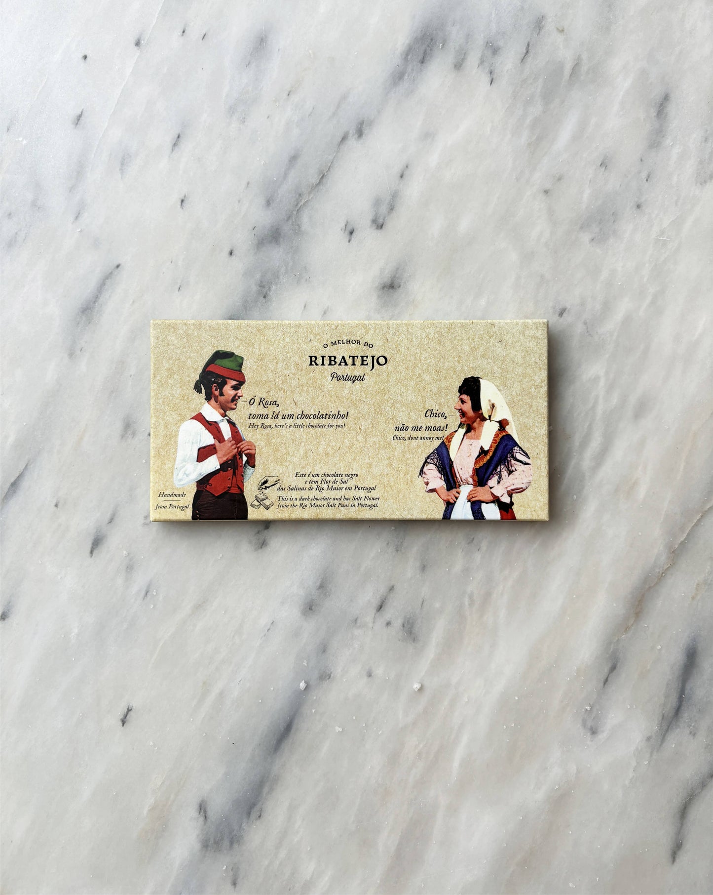 A rectangular package of O Melhor do Ribatejo Dark Chocolate with Flor de Sal, featuring a man and woman in traditional Portuguese clothing and Portuguese text on a beige background, rests on a white marble surface.