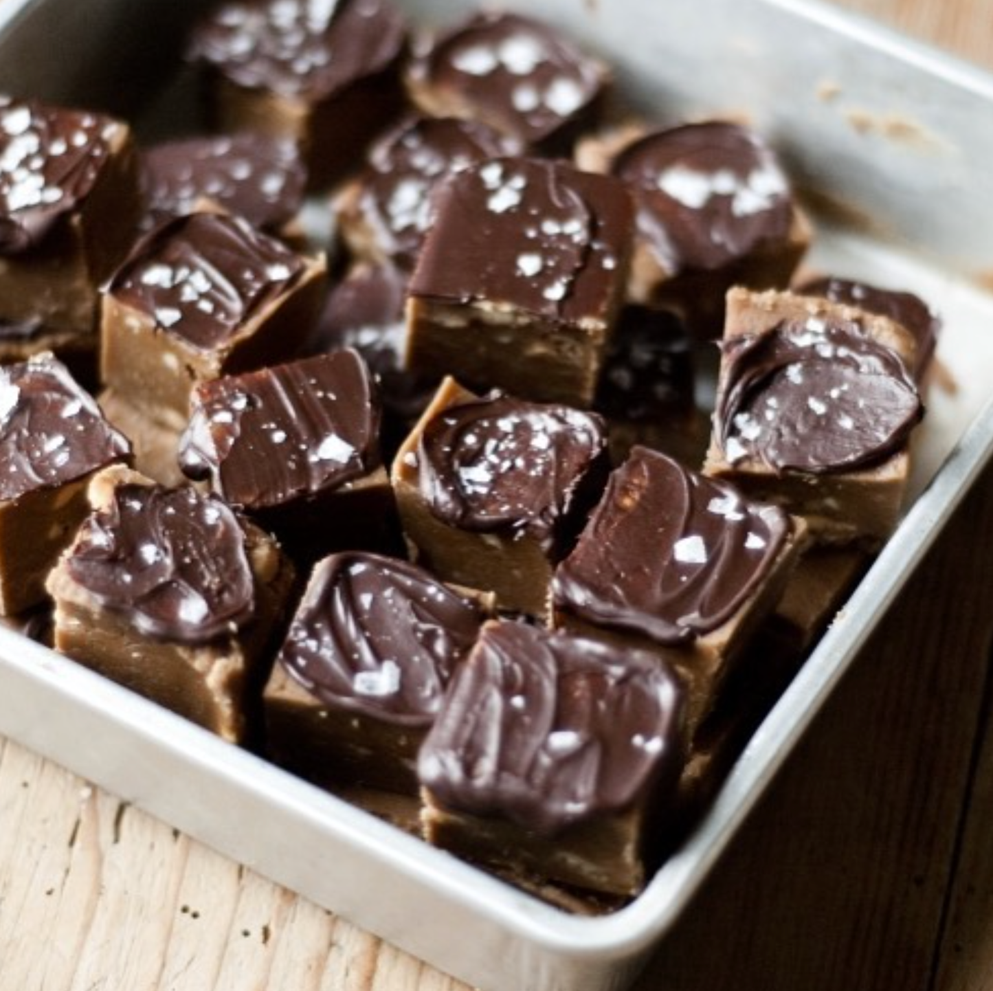 A metal baking pan filled with square chocolate fudge pieces topped with O Melhor do Ribatejo Salt Flower from Portugal, arranged closely together on a wooden surface.