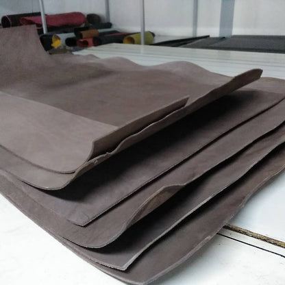 A Handcrafted Nubuck Leather Belt 3.5 cm by Creaciones Corpas is shown in a workshop, with flat sheets of brown Nubuck leather and rolled rubber sheets visible in the background.