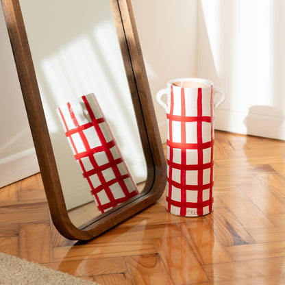 The Udine vase by OITO Atelier, a tall handmade ceramic piece with a red checkered pattern and two handles, stands on a wooden floor beside a wooden-framed mirror reflecting its artisanal charm in soft sunlight.