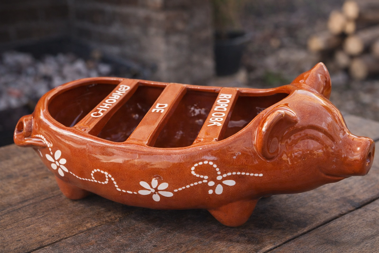 The Chorizo Pig Griller Dish by Ceramica Picas is a brown, pig-shaped dish with white floral patterns and labeled sections—Chicharrón, De, and Borrego—resting on a wooden surface outdoors.