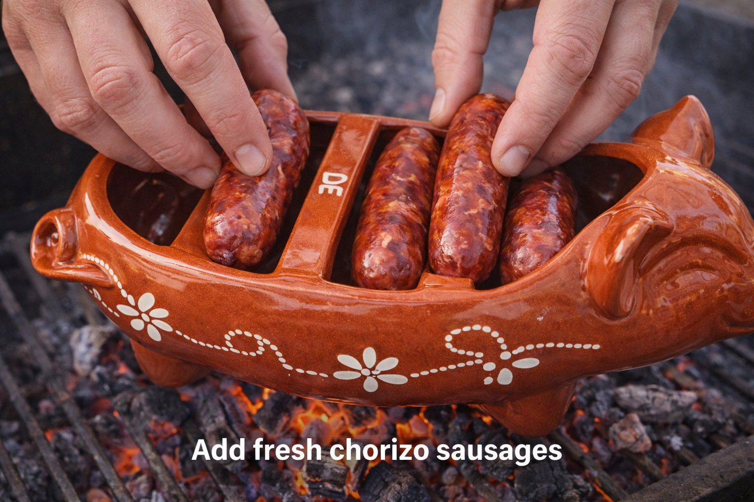 Two hands place smoked sausage into the Ceramica Picas Chorizo Pig Griller Dish over a fire, with "Add fresh chorizo sausages" text below.