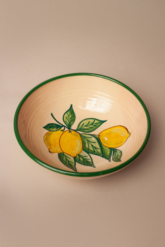 The Huerta Patterned Ceramic Salad Bowl by Eturel Madrid is a cream-colored bowl with a green rim, showcasing hand-painted yellow lemons and green leaves in a nature-inspired design inside.