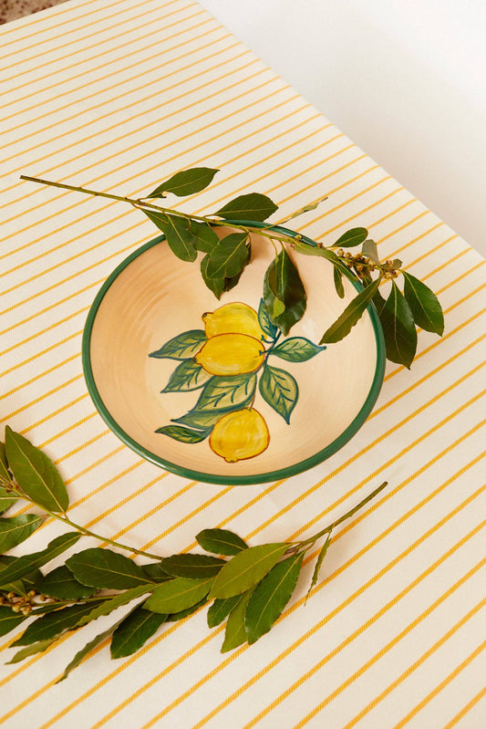 The Huerta Patterned Ceramic Salad Bowl by Eturel Madrid, featuring a nature-inspired lemon and leaf design, rests on a white tablecloth with yellow stripes, accented by green leaf sprigs.
