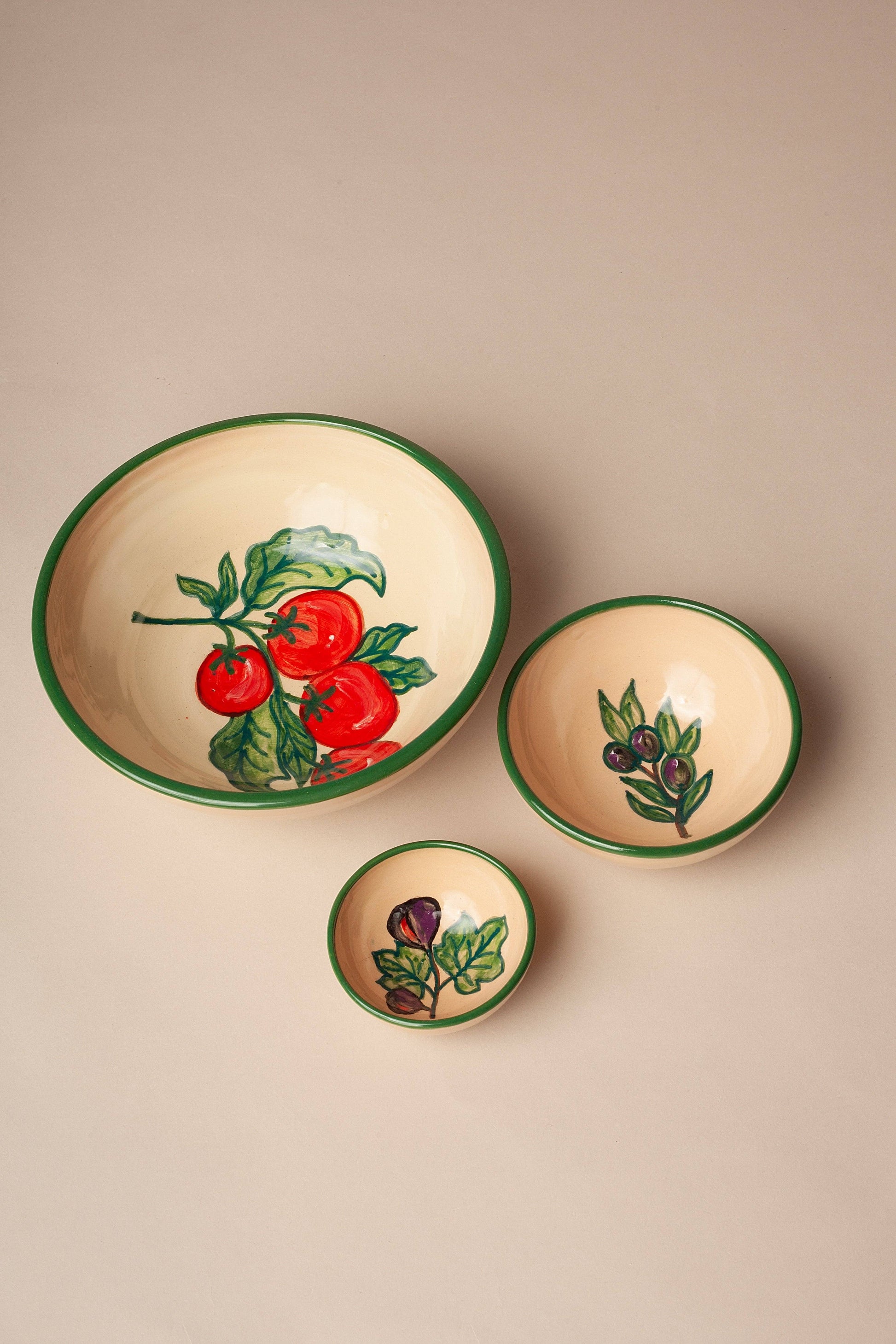 Three cream-colored Huerta Stamped Ceramic Bowls by Eturel Madrid, each with green rims and hand-painted tomato, olive, or fig designs, are arranged on a beige surface, showcasing handmade Spanish artisanal charm.