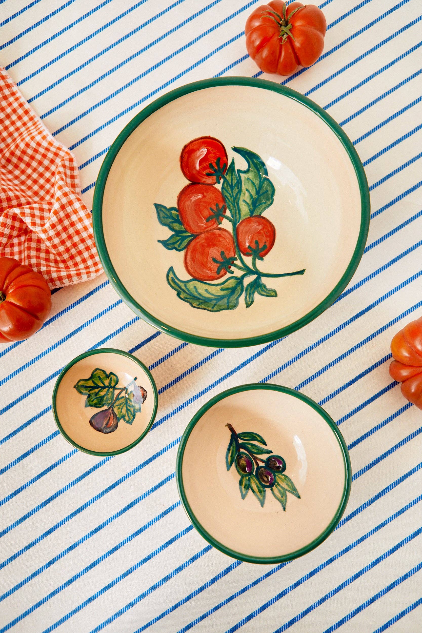 Three Eturel Madrid Huerta Stamped Ceramic Bowls, handmade in Spain with painted fruit motifs, rest on a blue-striped tablecloth amid tomatoes and orange gingham, exuding rustic artisanal charm.