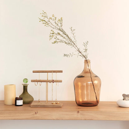 A wooden shelf holds a yellow candle, green vase, small jar, jewelry stand with necklaces and bracelets, a large Hannun Luiza Demijohn Vase with a sprig, and a white ceramic dish—showcasing sustainable design.