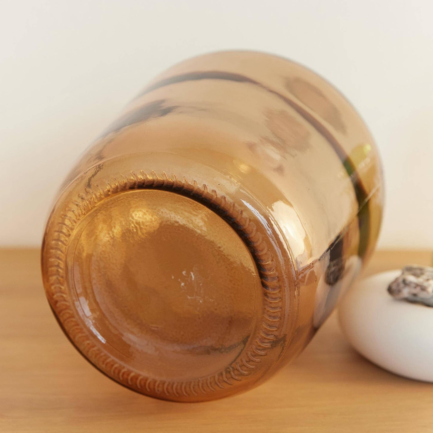 The Hannun Luiza Demijohn Vase, with its large brownish form and rounded base, rests on a wooden surface beside a small white item topped with a stone, embodying sustainable production and timeless design.