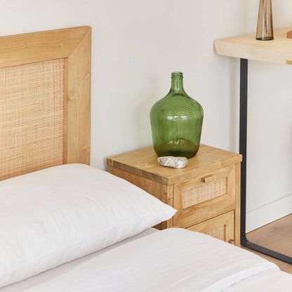 A neatly made bed with white linens sits beside a nightstand featuring the Luiza Demijohn Vase by Hannun, crafted sustainably. A wooden desk with a black leg appears in the background.