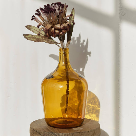 A single dried flower with dark tips rests in the Luiza Demijohn Vase by Hannun, placed on a round wooden stool and casting a soft shadow against a sunlit white wall.
