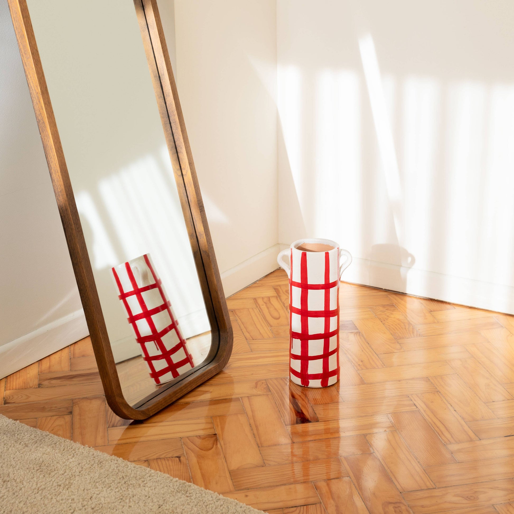 The Udine vase by OITO Atelier, featuring a handmade red grid pattern, sits on a wooden floor beside a large leaning mirror. Sunlight casts striped shadows, highlighting the artistry of this unique ceramic decor piece.