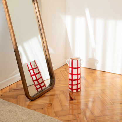 The Udine vase by OITO Atelier, featuring a handmade red grid pattern, sits on a wooden floor beside a large leaning mirror. Sunlight casts striped shadows, highlighting the artistry of this unique ceramic decor piece.