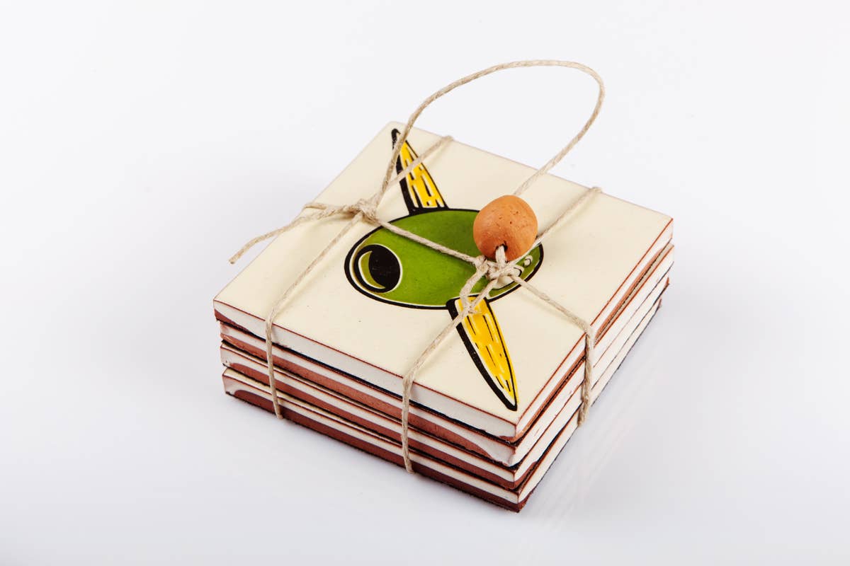 A set of four Bussoga Olivas Coasters, featuring a green and yellow abstract bird on white ceramic, are screen-printed and tied with string and a wooden bead.