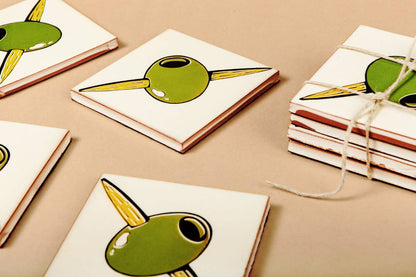 Several Bussoga Olivas Coasters, featuring charming green olives on toothpicks, are stacked and tied with string on a tan surface; these are screen-printed ceramic coasters.