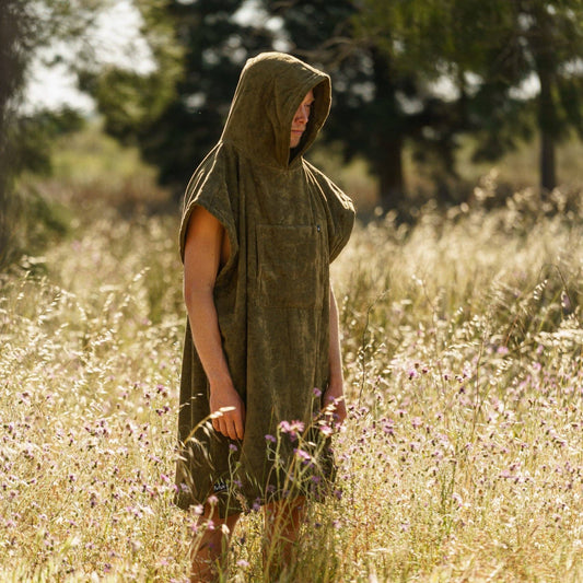 A person wearing the Futah Ericeira Terry Poncho, a sleeveless olive-green cotton hooded piece, stands in a sunlit field of tall grass and wildflowers with blurred trees in the background.