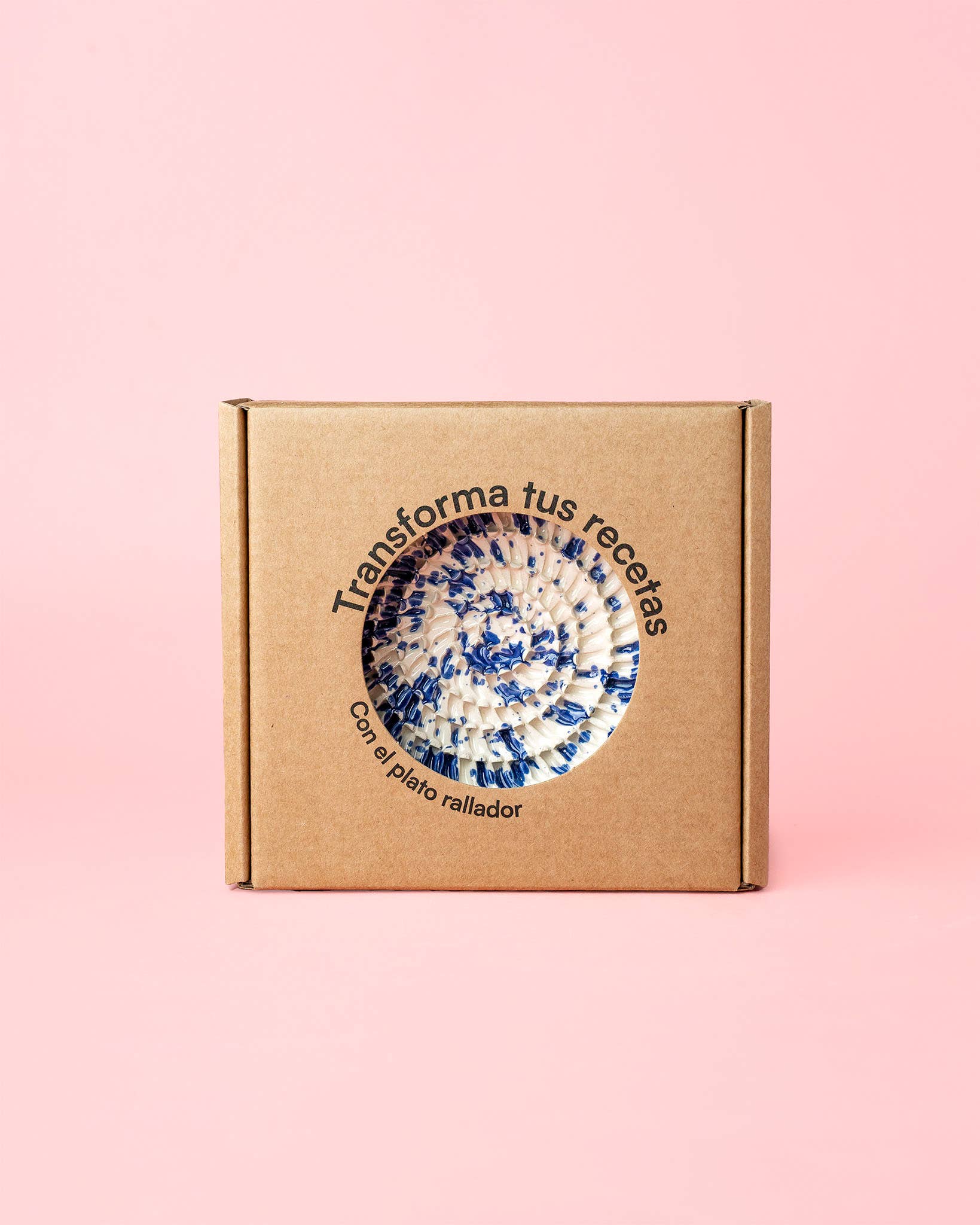 A brown cardboard box with a circular window showcases a blue and white Handmade Ceramic Grater Plate Small - Alcazaba by Alfar Tierra Cocida. Spanish text on pink reads: Transforma tus recetas con el plato rallador—an ideal kitchen gift.