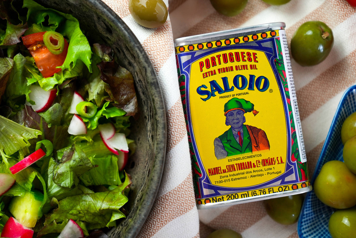 A 200ml can of Machado Saloio Olive Oil from Portugal sits on a striped cloth next to a bowl of fresh salad and green olives. The vibrant can features traditional Portuguese attire, emphasizing the brand’s heritage.
