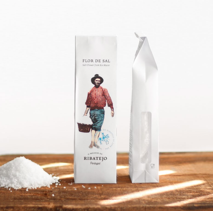 A box of O Melhor do Ribatejo's Salt Flower | From Portugal, featuring artwork of a basket-carrying person, rests on wood next to a small pile of this prized Portuguese salt from Ribatejo.