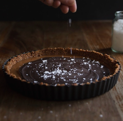 A hand sprinkles O Melhor do Ribatejo’s Salt Flower | From Portugal onto a chocolate tart with a graham cracker crust on a wooden table. A glass jar of this delicate sea salt sits in the background.