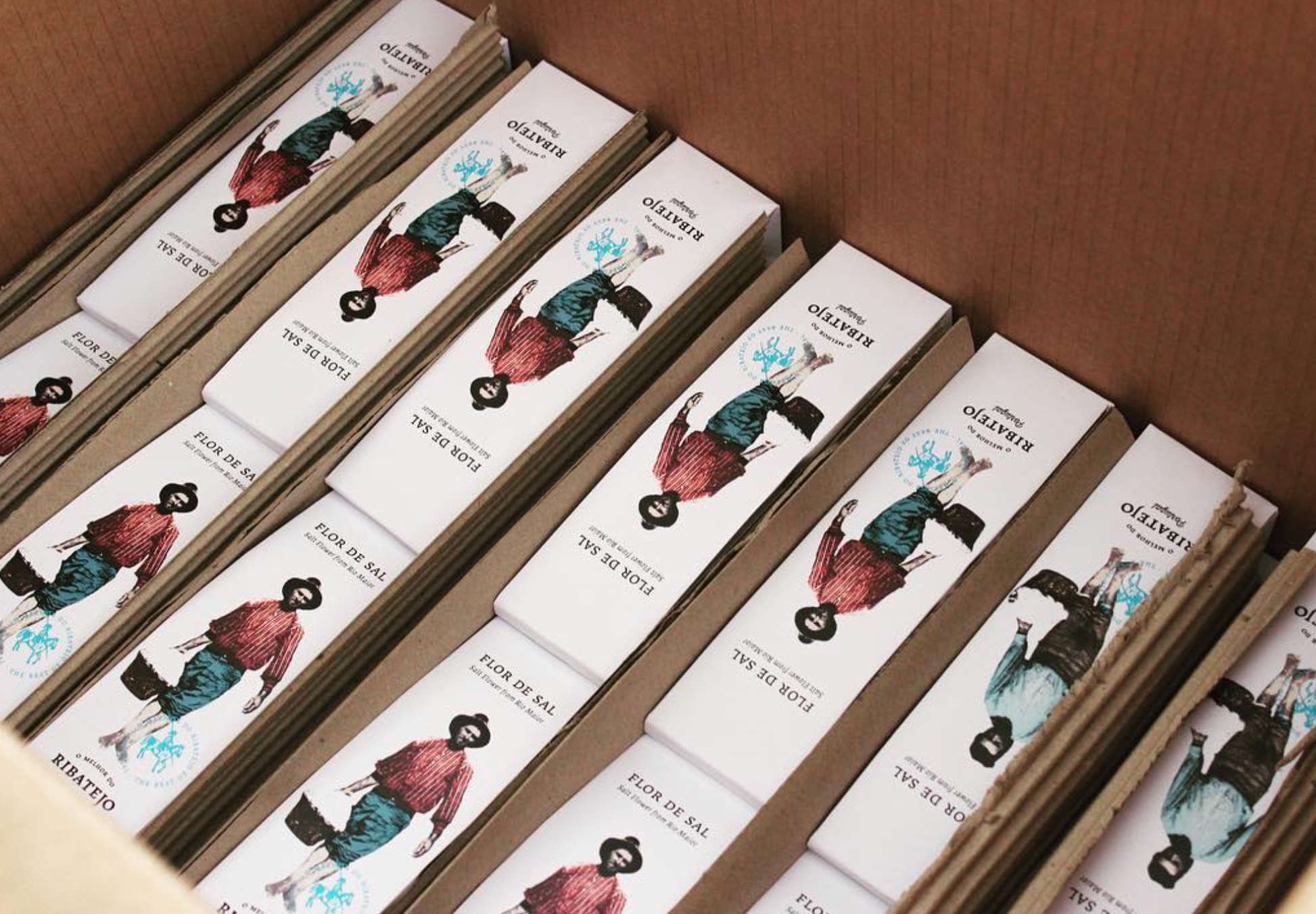 A cardboard box holds several rows of O Melhor do Ribatejo Salt Flower from Portugal, each package illustrated with a person in a hat, red shirt, and blue skirt. The boxes are arranged neatly inside.