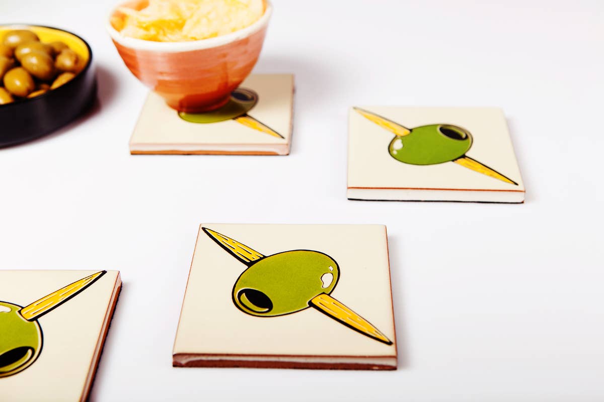 Four Bussoga Olivas Coasters, screen-printed with green olives on toothpicks, are arranged on a white surface. In the background, a bowl of green olives and another with yellow dip are partly visible.