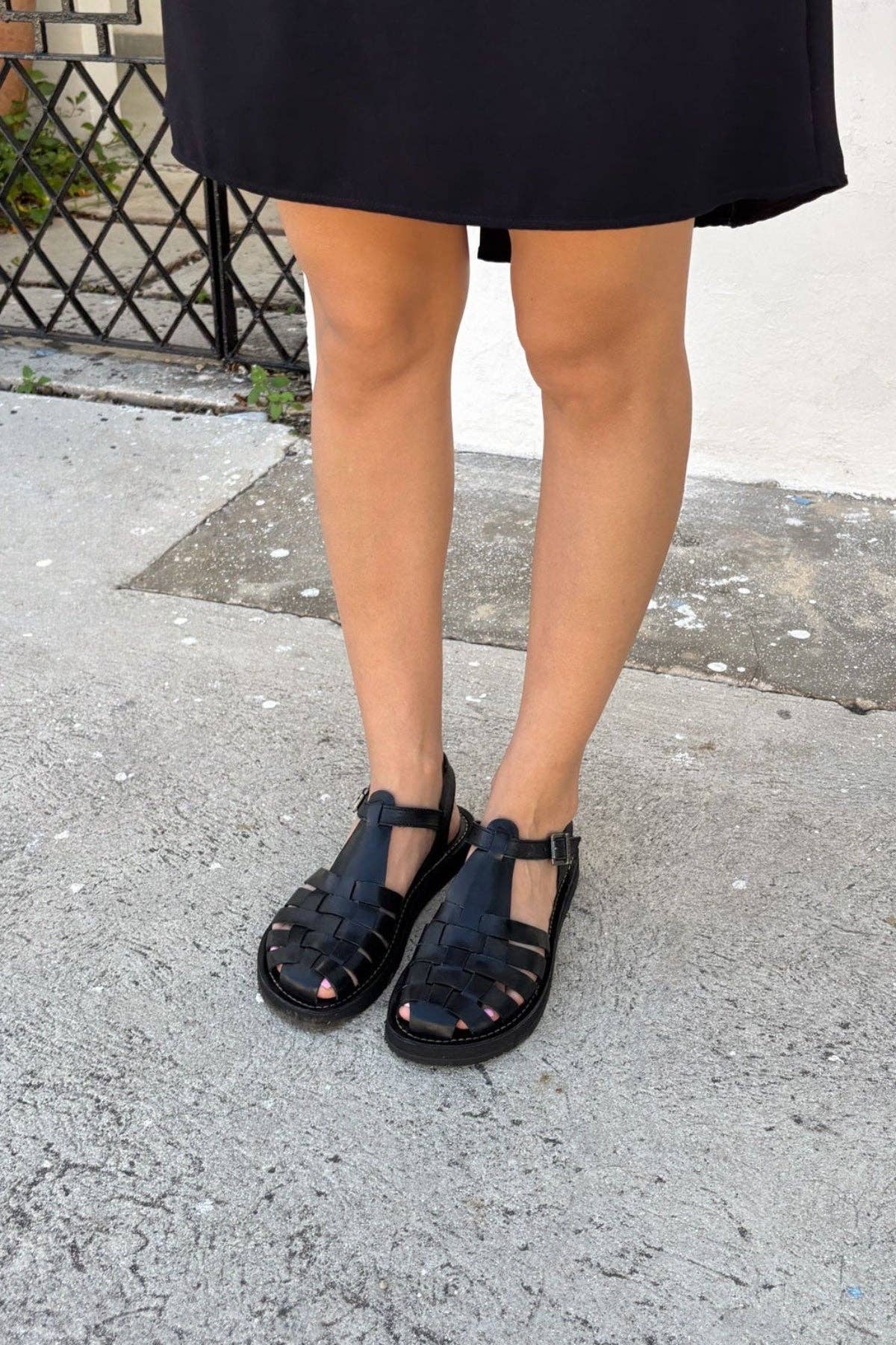 A person in a black knee-length skirt wears Mexas EU Black Glory sandals, standing on concrete near a white wall and metal gate. The stylish sandals elevate the outfit, adding refined elegance for any occasion.
