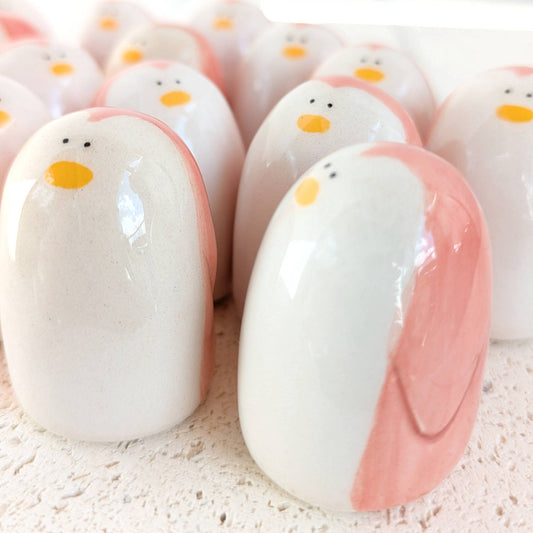 A cluster of Picuru ceramicas’ Penguin Amulets of Presence—handmade ceramic figurines with white, pink, and yellow details—rest on a textured surface, each symbolizing balance and resilience.