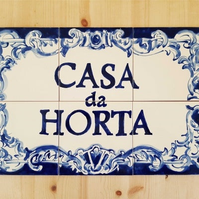 The Bespoke Outdoor House Address Portuguese Tile Mural in Cobalt Blue by TilePassion features ornate cobalt blue designs framing bold lettering on a white ceramic tile, elegantly mounted on a light wooden surface.