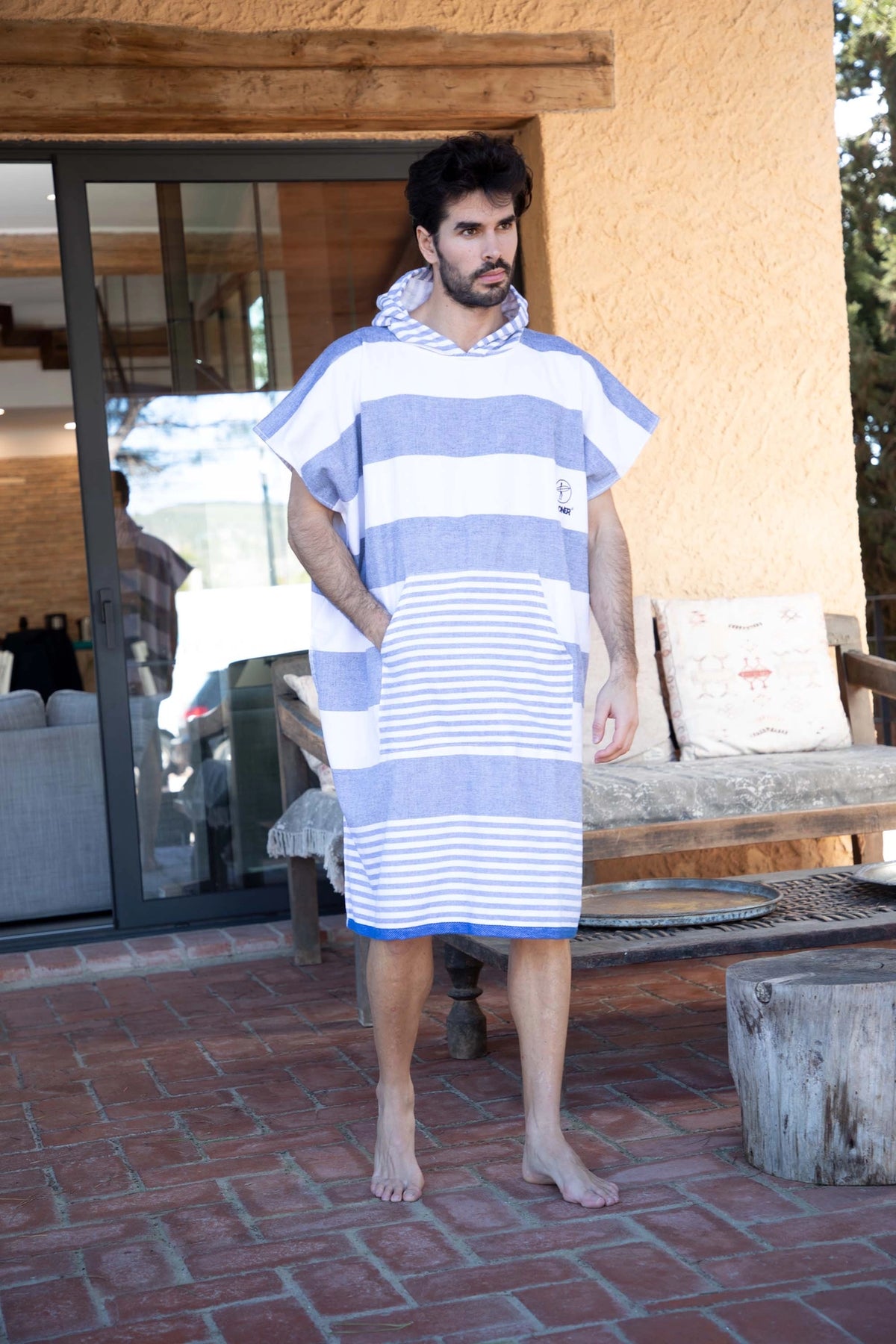 A man wearing a blue and white striped hooded towel cover-up robe, standing barefoot on a brick patio in front of a modern house.