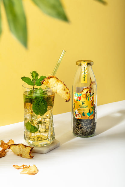 A glass of iced tea with mint, a glass straw, and a dried pineapple garnish sits beside a bottle of Criatura Organic Mojito Cocktail Infusion – Pineapple, Apple & Mint. Dried pineapple pieces are scattered on a white surface with a yellow background.