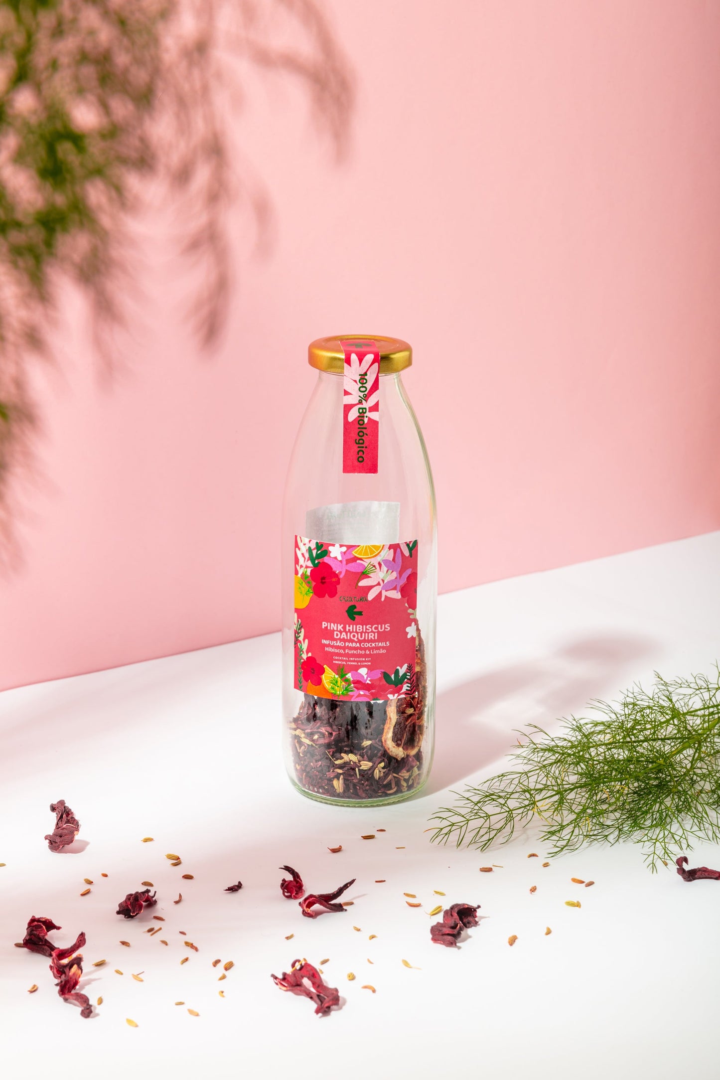 A glass bottle of Criatura Pink Hibiscus Daiquiri, with a gold cap and pink floral label, filled with dried hibiscus flowers, rests on a white surface against a pink background, surrounded by scattered petals and leafy sprigs.