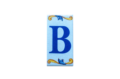 A Lisa Croquets Letters – Ceramic House Letter Tiles rectangular tile featuring a blue letter B centered between yellow and blue scrolls on a light blue crackle-finish background.